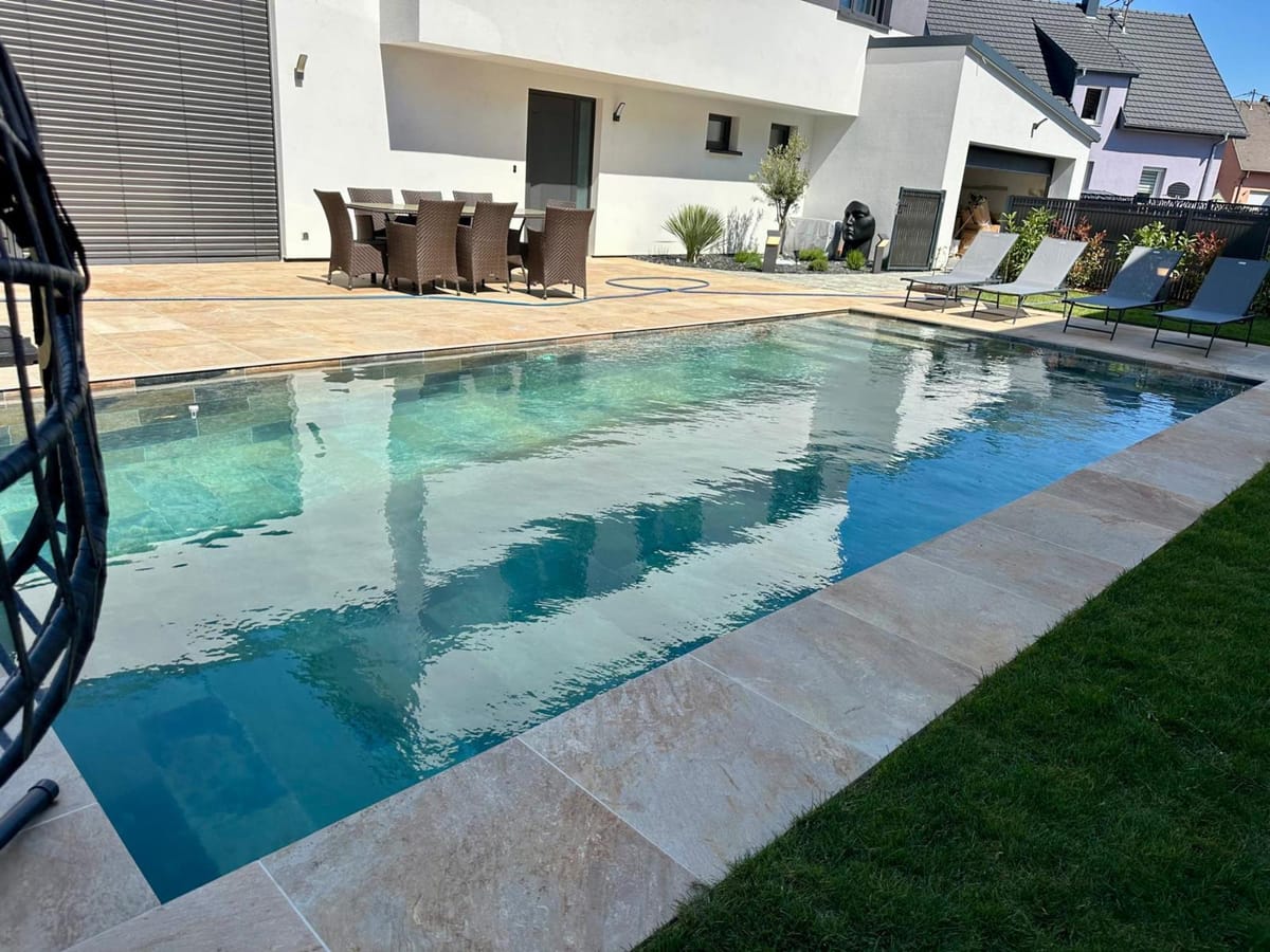 Piscine longue devant une maison contemporaine, margelles et plage en pierre reconstituée