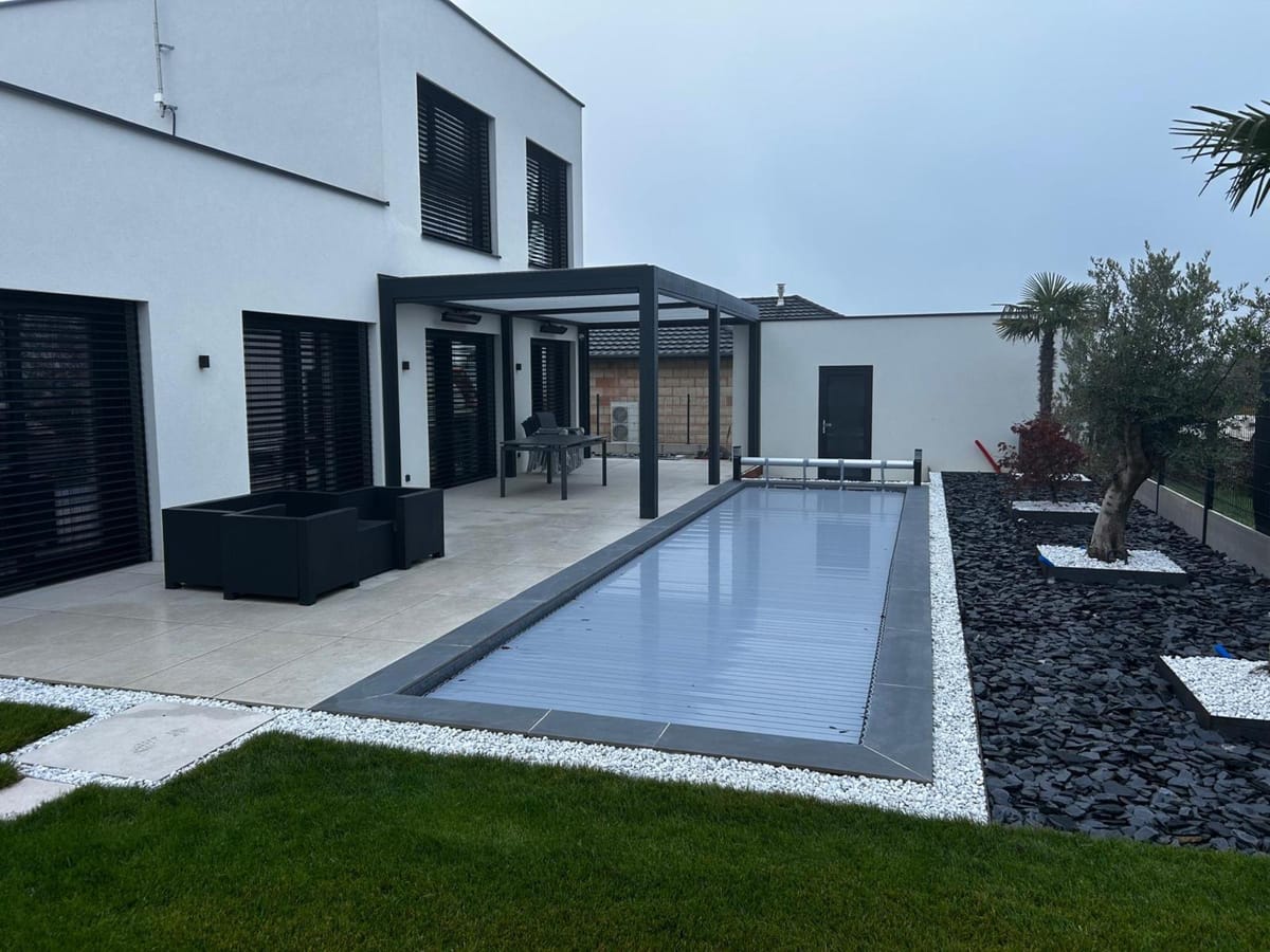 Piscine à volet immergé devant une maison moderne, plage en dalles et massif de graviers